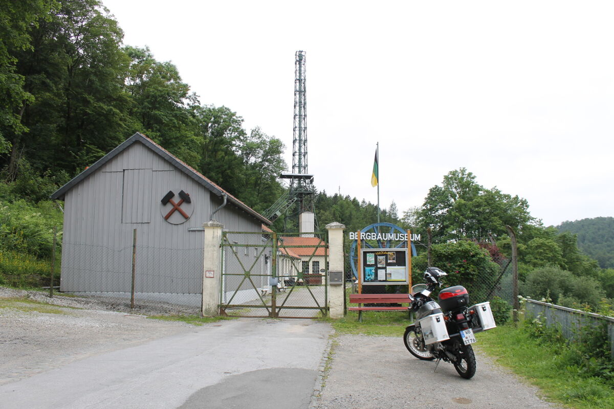 Graue Werkshalle mit Schlägel und Eisen am Giebel, dahinter ein stählernes Fördergerüst, davor ein abgestelltes Motorrad am Tor Graue Werkshalle mit Schlägel und Eisen am Giebel, dahinter ein stählernes Fördergerüst, davor ein abgestelltes Motorrad am Tor